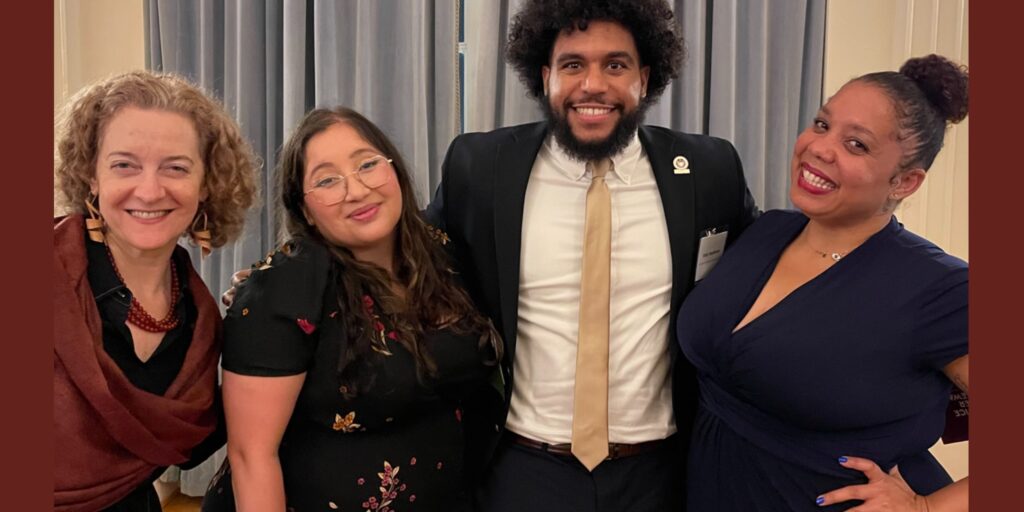 Four adults of different ethnicities dressed formally standing in a group and smiling at the camera.