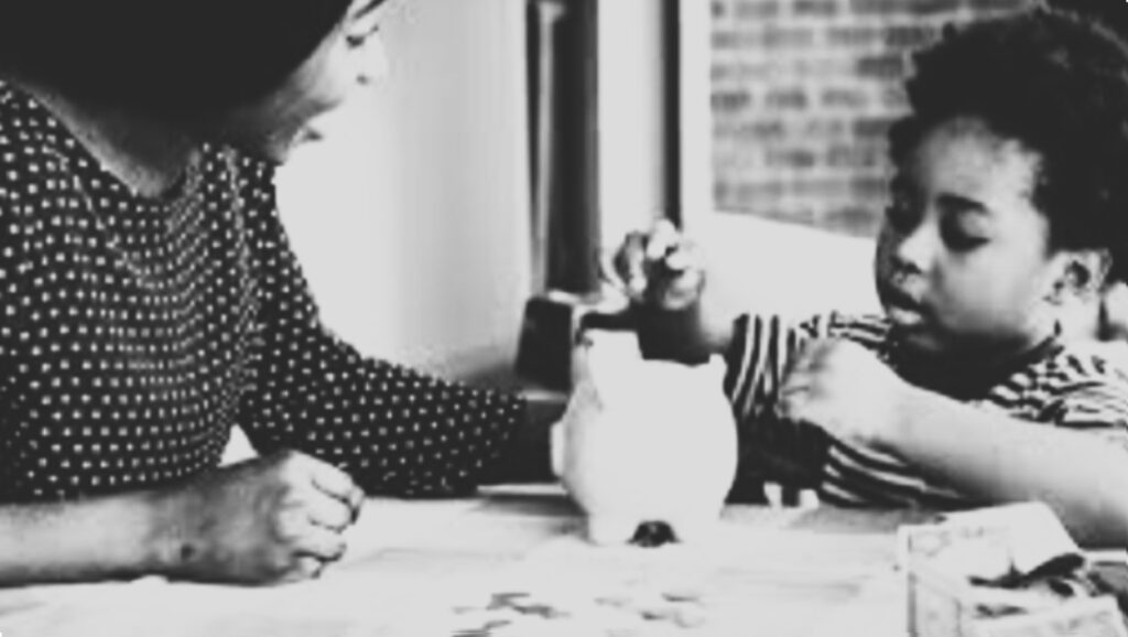 A woman watching a young child put money in a piggy bank.